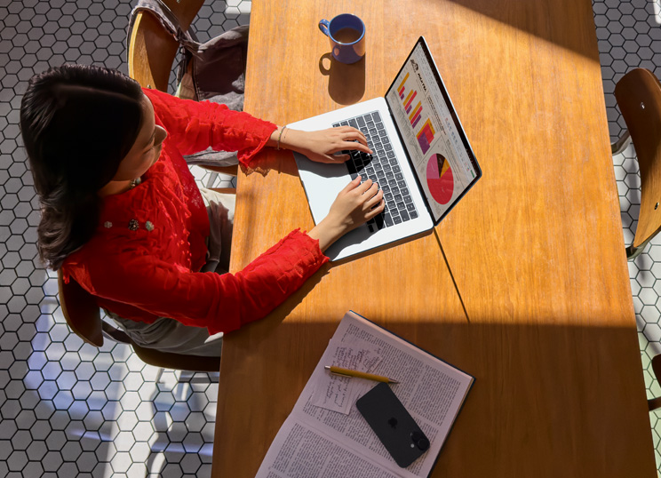 Una persona usa una MacBook en una mesa, con una taza, un smartphone y papeles cerca.